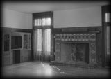 Irving Gill's Dodge House, a mid-century modern house. Black and white photo of the living room with fireplace surrounded by tile, tall windows on either side.